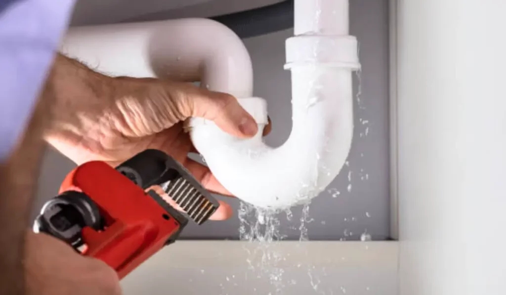 A professional plumber is performing a pipe repair on a leaking white PVC pipe under a sink, using a wrench to tighten the fittings.