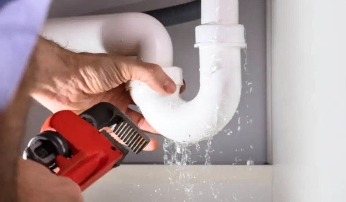 A professional plumber is performing a pipe repair on a leaking white PVC pipe under a sink, using a wrench to tighten the fittings.