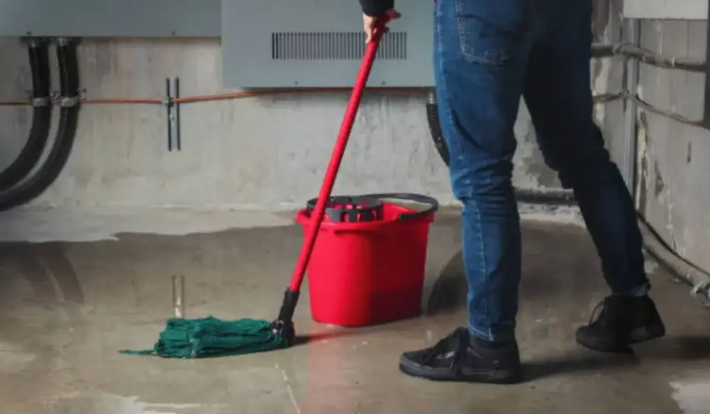 Person mopping a flooded basement with a red bucket, cleaning water damage caused by plumbing issues or heavy rain.