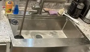 Stainless steel kitchen sink with soap scum, a plunger, and cleaning tools, showing the need for drain cleaning.