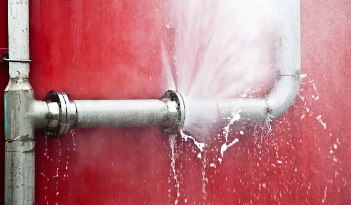 Metal pipe bursting due to high water pressure, spraying water forcefully against a red industrial wall.