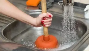 A person using a plunger on a backed-up kitchen sink while water runs, showing a common DIY drain cleaning attempt.