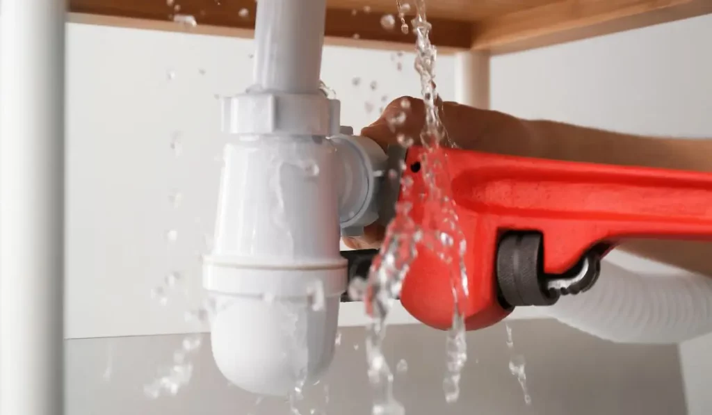 Water leaking from a white PVC pipe under a sink while a plumber uses a red pipe wrench to perform emergency service in a Naperville home.