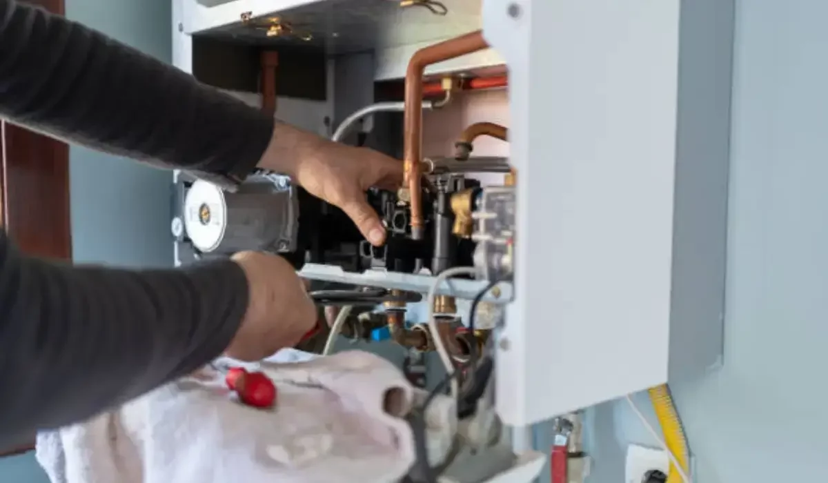 Technician repairing a broken winter water heater, adjusting copper pipes and wiring inside utility room unit.